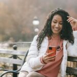 A cheerful woman enjoying a sunny autumn day, sitting on a bench and browsing her smartphone in the park.